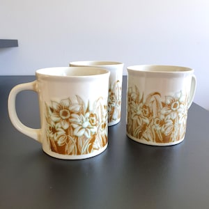 May include: Three ceramic mugs with a cream-colored base and a floral design in shades of brown and light green. Each mug has a handle and a cylindrical shape, perfect for enjoying hot beverages.