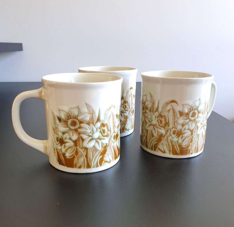 May include: Three ceramic mugs with a cream-colored base and a floral design in shades of brown and light green. Each mug has a handle and a cylindrical shape, perfect for enjoying hot beverages.