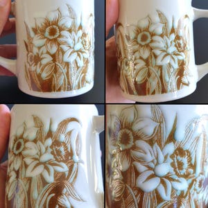 May include: White ceramic mug featuring a floral design in shades of brown and light blue. The flowers and leaves are embossed on the mug's surface. The mug has a curved handle and a slightly flared rim.