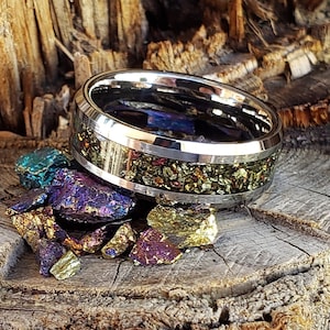 May include: A silver ring with a band of iridescent pyrite embedded in epoxy resin. The ring is sitting on a piece of wood with small pieces of pyrite scattered around it.