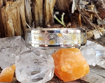 Puede incluir: Un anillo de plata con una incrustación de resina transparente que contiene pequeñas motas doradas. El anillo está apoyado en una pieza de madera con varios cristales de cuarzo transparentes y un pequeño cristal naranja.
