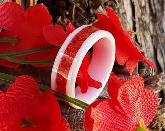 Puede incluir: Un anillo blanco con una incrustación de purpurina roja. El anillo está sobre una superficie de madera con flores rojas.