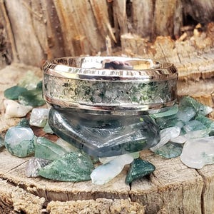 May include: A silver ring with a wide band featuring a moss agate inlay. The ring is set on a bed of green and clear gemstones, resting on a weathered wooden surface. The ring's design showcases a natural, earthy aesthetic.