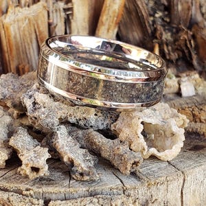 May include: A silver-toned ring with a dark, textured center band. The ring is set against a backdrop of rough, light-colored organic material and a weathered wooden surface, creating a rustic aesthetic. The ring's design is simple yet striking.
