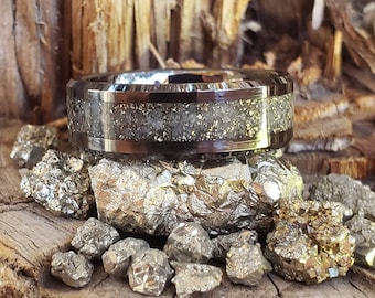 Puede incluir: Un anillo de carburo de tungsteno plateado con una incrustación negra y un acabado plateado moteado. El anillo está sentado sobre un lecho de pequeñas piedras brillantes de color plateado.