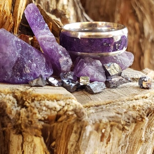 May include: A silver ring with a purple inlay, surrounded by amethyst crystals and other purple stones on a wooden surface.