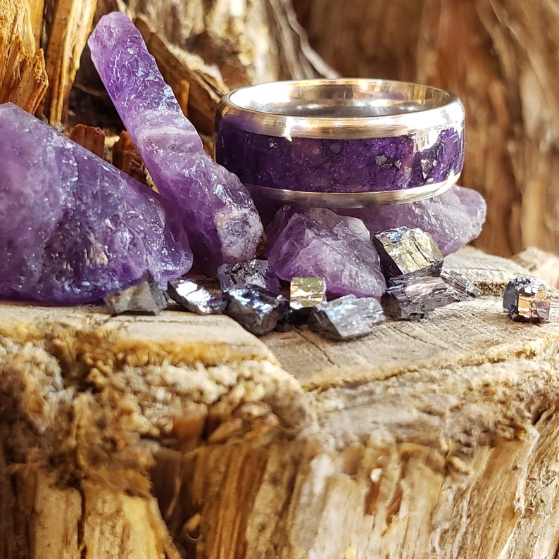 May include: A silver ring with a purple inlay, surrounded by amethyst crystals and other purple stones on a wooden surface.