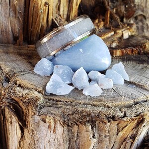 Może przedstawiać: Srebrny pierścionek z białym wkładką leży na jasnoniebieskim krysztale selenitu. Kryształ jest otoczony mniejszymi kryształami selenitu na drewnianej powierzchni.