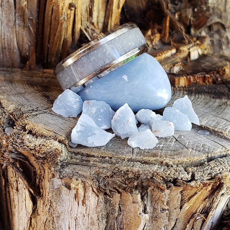 Może przedstawiać: Srebrny pierścionek z białym wkładką leży na jasnoniebieskim krysztale selenitu. Kryształ jest otoczony mniejszymi kryształami selenitu na drewnianej powierzchni.