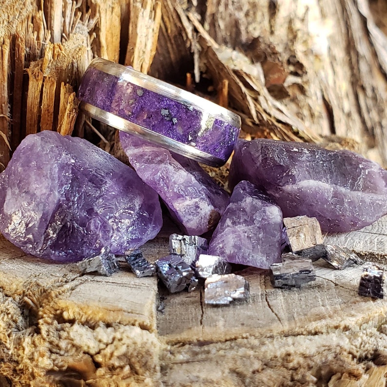 May include: A silver ring with purple amethyst stones embedded in the band. The ring is surrounded by small amethyst crystals and a few metallic silver crystals.