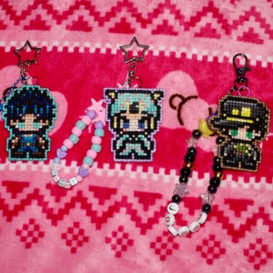 May include: Three pixel-art style keychains featuring embroidered character designs. The left keychain has blue hair, the center has teal hair, and the right has a black hat. Two keychains include beaded loops with letter beads spelling names.