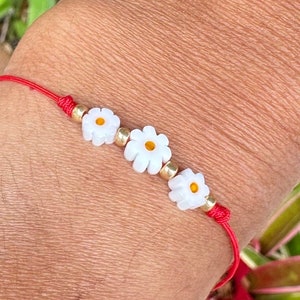 May include: A red string bracelet with three white daisy-shaped beads with yellow centers.