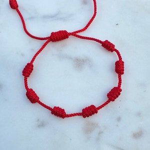 May include: A red string bracelet with knotted beads. The bracelet is on a white marble surface.