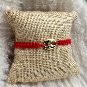 May include: Red braided bracelet with a gold-toned anchor charm. The bracelet is displayed on a small burlap pillow.