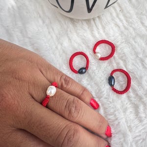 May include: Four red string rings with a pearl and black bead accent. One ring is worn on a finger.