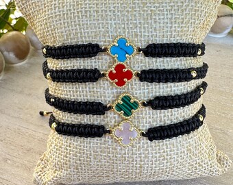 May include: Four black braided bracelets with gold clover charms. Each charm has a different colored stone: turquoise, red, green, and pink.
