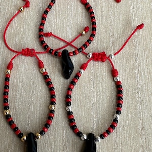 May include: Three adjustable bracelets with red cords, black and red beads, and a black pendant. The bracelets have gold and silver accent beads. The bracelets are displayed on a light wood surface.