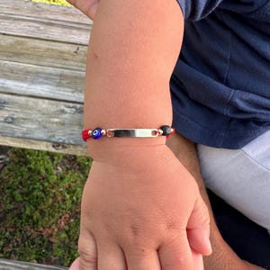 Puede incluir: Una pulsera roja y dorada con una placa de identificación plateada, cuentas azules y negras y un amuleto de ojo turco. La pulsera se lleva en la muñeca de un niño.