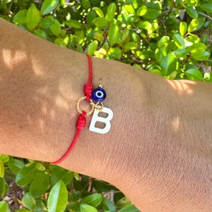 Puede incluir: Pulsera de hilo rojo con un dije de letra "B" blanca y una cuenta azul de ojo turco. La pulsera tiene detalles dorados y se encuentra sobre un fondo de follaje verde.
