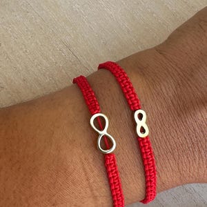 Peut inclure: Deux bracelets rouges en ficelle, ornés de breloques dorées en forme de symbole infini et du chiffre huit. Les bracelets sont portés sur un poignet, mettant en valeur le rouge vif de la ficelle et l'éclat métallique des breloques. Le fond est une surface en bois clair.