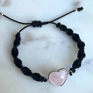 May include: Black macrame bracelet featuring a pink heart-shaped gemstone set in a silver-colored bezel. The bracelet is adjustable with a sliding knot closure and small silver beads.