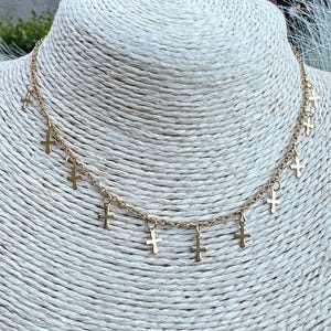 May include: Gold-coloured necklace with multiple small cross pendants. The necklace is displayed on a cream-coloured bust, with a blurred green background. The chain and crosses appear to be made of a shiny metal.