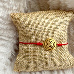 Pode incluir: Um bracelete de corda vermelha com um amuleto de espiral dourado. O bracelete está sobre um travesseiro de juta.