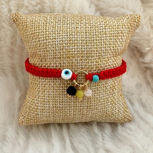 May include: Red string bracelet with a blue evil eye charm and gold beads. The bracelet has three additional charms: a black bead, a light green bead, and a pink bead.
