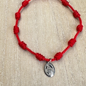 Peut inclure: Bracelet en cordon rouge noué avec un pendentif argenté. Le bracelet présente plusieurs nœuds le long du cordon et un petit pendentif rond avec l'image d'une figure religieuse. Le bracelet est présenté sur une surface en bois clair.