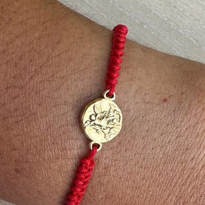 Könnte beinhalten: Ein rotes geflochtenes Armband mit einem goldfarbenen kreisförmigen Anhänger. Der Anhänger hat ein eingraviertes Design. Das Armband wird am Handgelenk getragen.