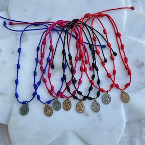 May include: A collection of colorful, knotted cord necklaces. Each necklace features a small, round pendant and is available in blue, pink, black, and red. The necklaces are displayed on a white, marble-like surface.