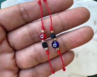 May include: Two red string bracelets featuring small beads. One bracelet has a red evil eye bead and a black cube bead. The other bracelet has a blue evil eye bead and a black cube bead. Both bracelets include small metallic spacer beads.