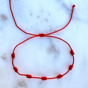 Puede incluir: Un brazalete de cuerda roja con múltiples nudos. El brazalete está sobre una superficie de mármol blanco.