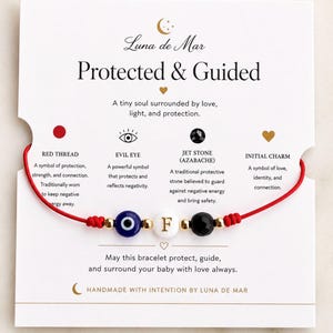 May include: A red string bracelet featuring a blue evil eye bead, a white bead with the letter F, and a black stone bead, accented with small gold-tone beads. The bracelet is displayed on a white card with the text Luna de Mar Protected & Guided.