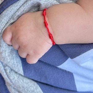 May include: A close-up of a baby's hand wearing a red string bracelet. The bracelet is tied in a knot and has a simple design.