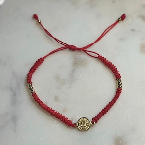 Könnte beinhalten: Rotes Makramee-Armband mit goldfarbenen Perlen und einem kleinen, runden goldfarbenen Anhänger. Das Armband ist mit einem Gleitknotenverschluss geknotet. Der Anhänger hat ein detailliertes Design. Das Armband wird auf einer weißen Marmoroberfläche präsentiert.