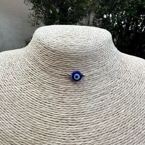 May include: A small, round, blue and white evil eye charm on a silver-colored metal pin. The charm is centered on a beige, textured surface, likely a display bust. The background features green foliage.