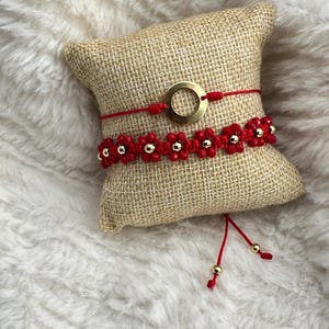 May include: Two red string bracelets with gold beads and a gold circle charm on a burlap cushion. The bracelets are tied together with a red string.