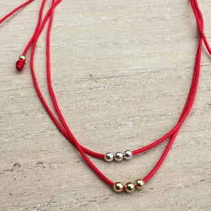 Puede incluir: Dos pulseras de hilo rojo con nudos ajustables. Una pulsera presenta tres cuentas plateadas, y la otra tiene tres cuentas doradas. Las pulseras se exhiben sobre una superficie de madera clara.