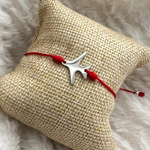 May include: Red string bracelet with a silver aeroplane charm. The bracelet is on a hessian cushion.