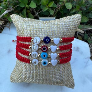 Puede incluir: Cuatro pulseras rojas tejidas con cuentas doradas que deletrean "LUNA", cuentas blancas en forma de corazón y cuentas azules de ojo turco. Las pulseras se exhiben sobre una almohada de arpillera beige, con follaje verde en el fondo.