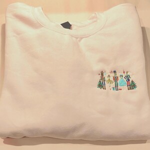 May include: White sweatshirt with a festive embroidered design featuring a nutcracker, a ballerina, and other characters. The design is in shades of green, red, blue, and gold.