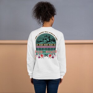 Puede incluir: Sudadera blanca con un estampado gráfico en color verde azulado y rosa en la parte posterior. El gráfico presenta una calavera con una corona, un diseño floral y el texto "...and a queen will always turn pain into power".