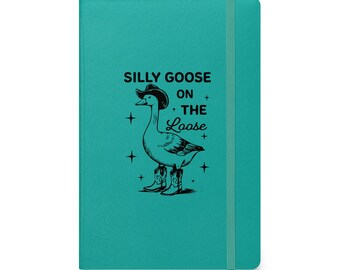 May include: A teal notebook with a black and white illustration of a goose wearing a cowboy hat and boots. The text "Silly Goose on the Loose" is above the illustration. A teal elastic band is on the right side.