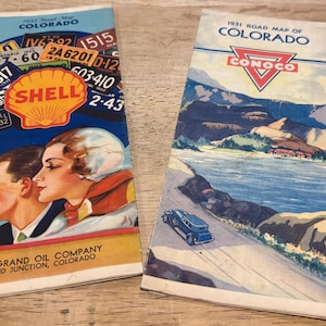 May include: Two vintage paper road maps of Colorado. The left map features a Shell logo, license plates, and a couple. The right map shows a Conoco logo, a mountain lake landscape, and a blue car. Both include text identifying the year and location.