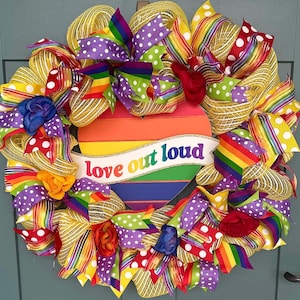 Op de afbeelding: Een levendige, handgemaakte krans met een regenbooghart en de tekst "love out loud". De krans is versierd met kleurrijke linten, strikken en gaas in regenboogkleuren, ter viering van de pride.