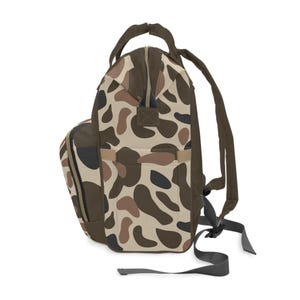 May include: A backpack with a camouflage pattern in shades of brown, tan, and black. The bag has a top handle, adjustable shoulder straps, and a side pocket. The zipper is brown.