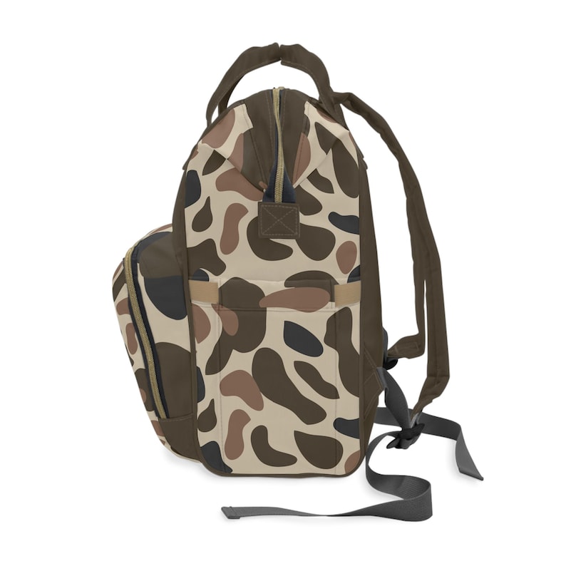 May include: A backpack with a camouflage pattern in shades of brown, tan, and black. The bag has a top handle, adjustable shoulder straps, and a side pocket. The zipper is brown.