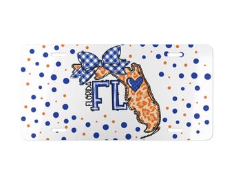 May include: A rectangular white license plate featuring a blue and orange polka dot pattern. The center displays a map of Florida with an orange leopard print, a blue and white gingham bow, a blue heart, and the text FLORIDA FL in blue letters.
