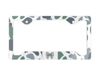 May include: A rectangular license plate frame featuring a white background with an abstract pattern of sage green, slate blue, and dark gray organic shapes. A small, detailed black and white leopard print bow is centered at the bottom edge.